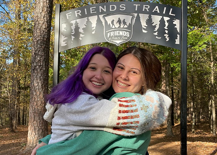 Two women embrace in front of a sign that reads 'Friends Trail'.