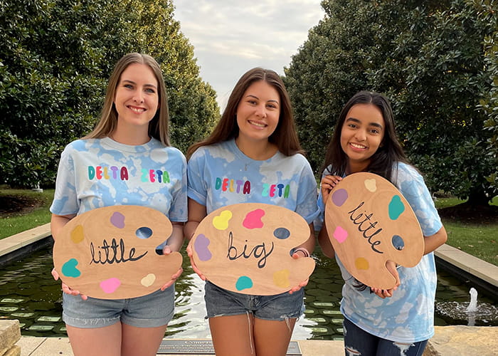 Three women in Delta Zeta shirts pose with signs displaying 'Little, Big, Little'.