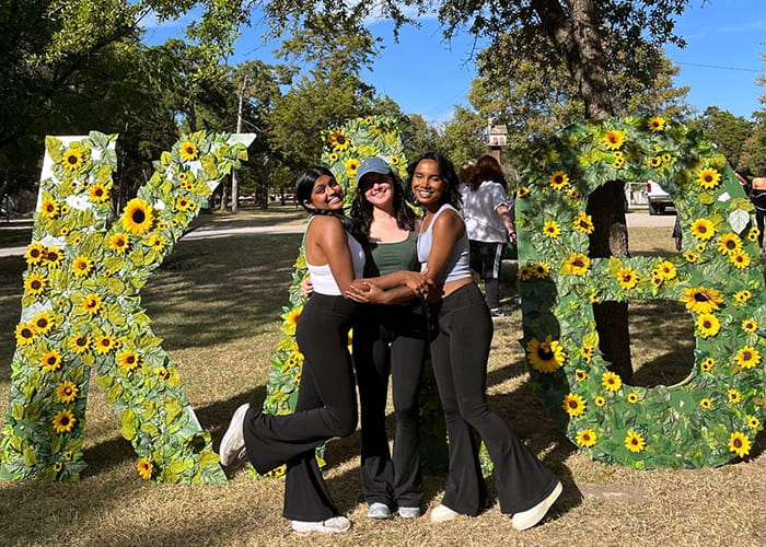 Three women pose in front of large greek letters Kappa Alpha Theta.