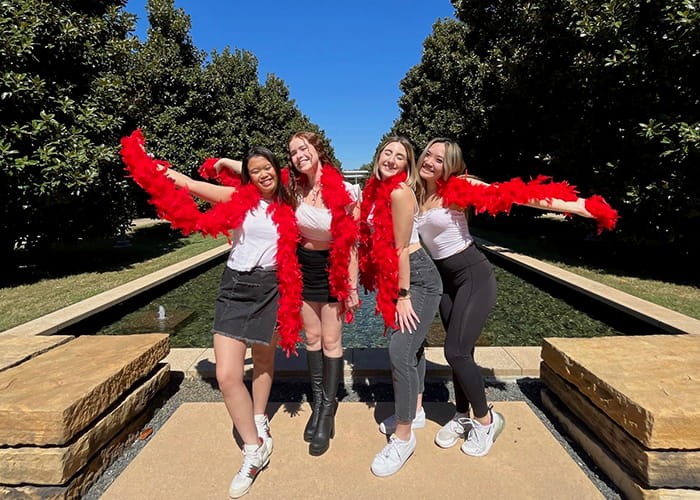 Four women with red feather boas pose at the reflecting pools at UT Dallas.