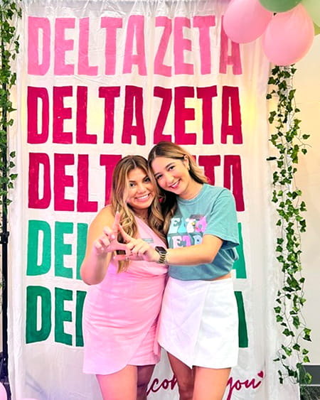 Two women pose in front of a backdrop with repeating Delta Zelta lettering.