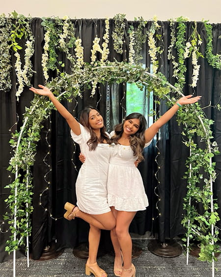 Two women with arms raised pose under a green archway.