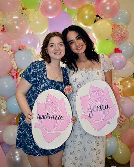 Two women pose with identifying signs against a backdrop of pastel colored balloons.